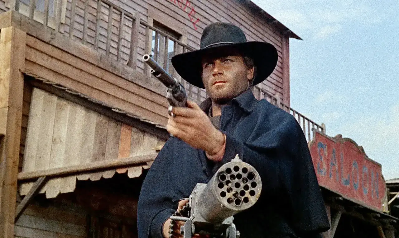 Franco Nero w kapeluszu i ciemnym płaszczu, z rewolwerem i karabinem Gatlinga, na tle drewnianego saloonu.