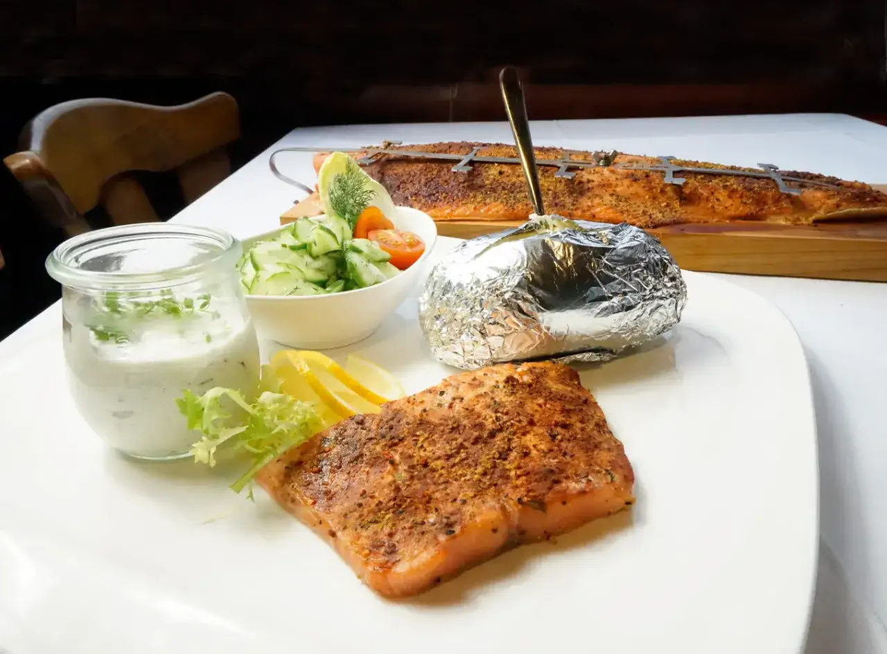 Flammlachs mit Beilagen: Ein saftiges Lachsfilet, Folienkartoffel, Gurkensalat und Dip. Im Hintergrund ein ganzer Flammlachs.
