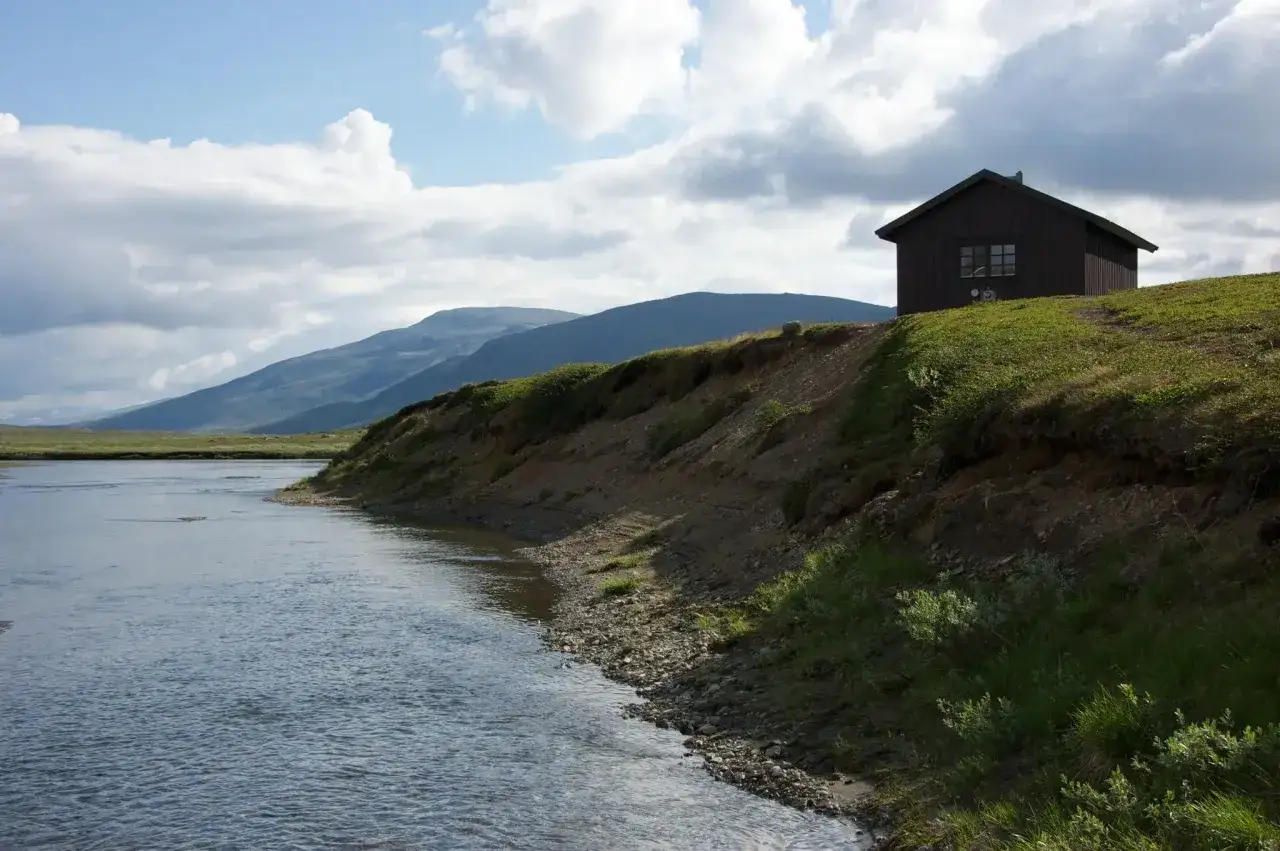 En m&ouml;rk stuga p&aring; en gr&ouml;n kulle vid en forsande &auml;lv i Padjelanta nationalpark. Berg i bakgrunden under en molnig himmel.
