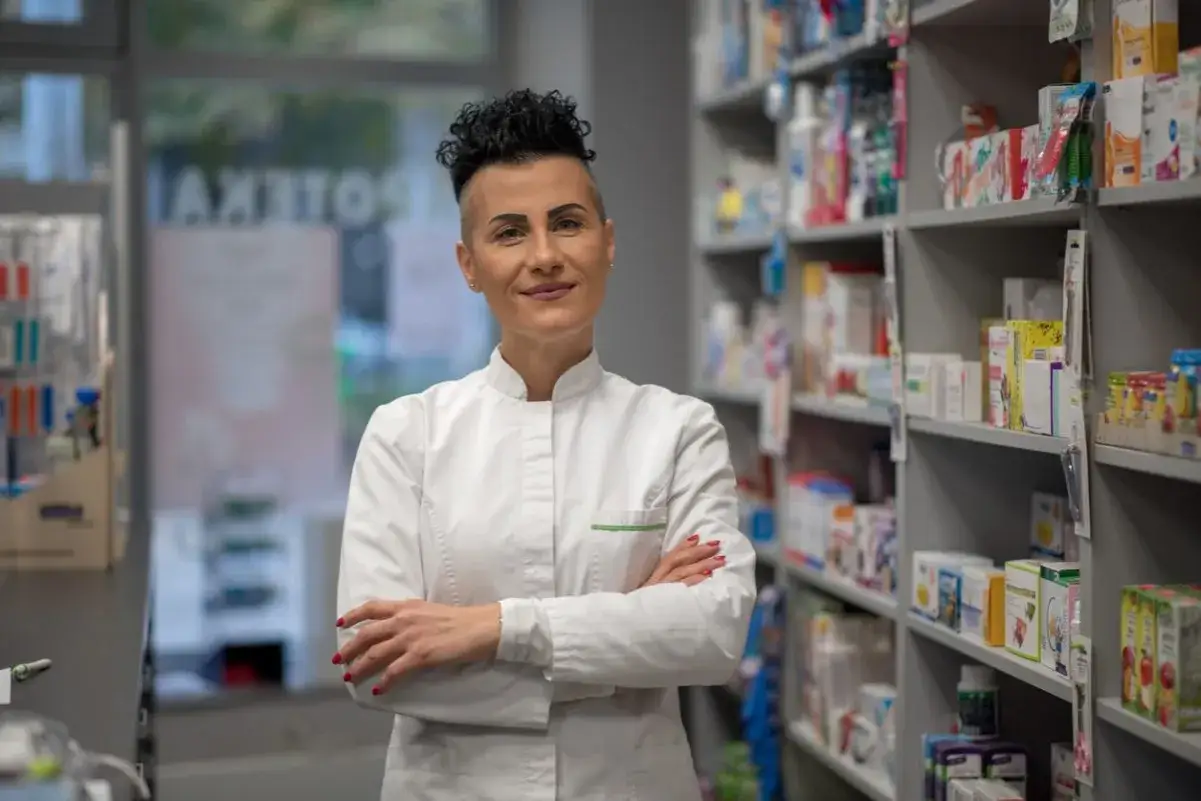 Uśmiechnięta farmaceutka w białym fartuchu stoi na tle aptecznych półek pełnych leków i kosmetyków.