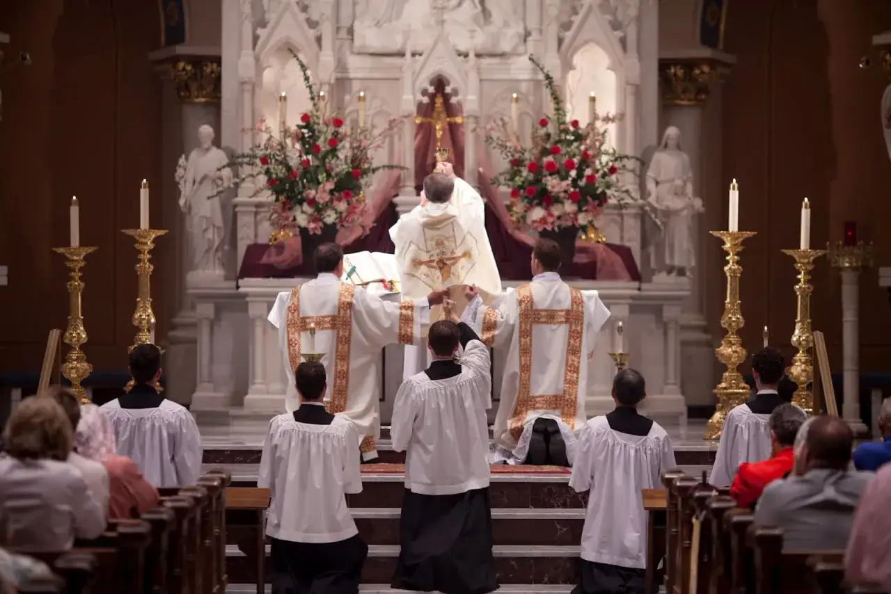 Uroczysta msza w kościele. Kapłani w szatach liturgicznych, wierni klęczący w ławkach.
