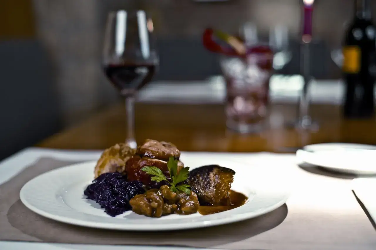 Ein Teller mit deftigem Essen zur Weinprobe: Fleisch, Rotkohl und Kl&ouml;&szlig;e, dazu ein Glas Rotwein.