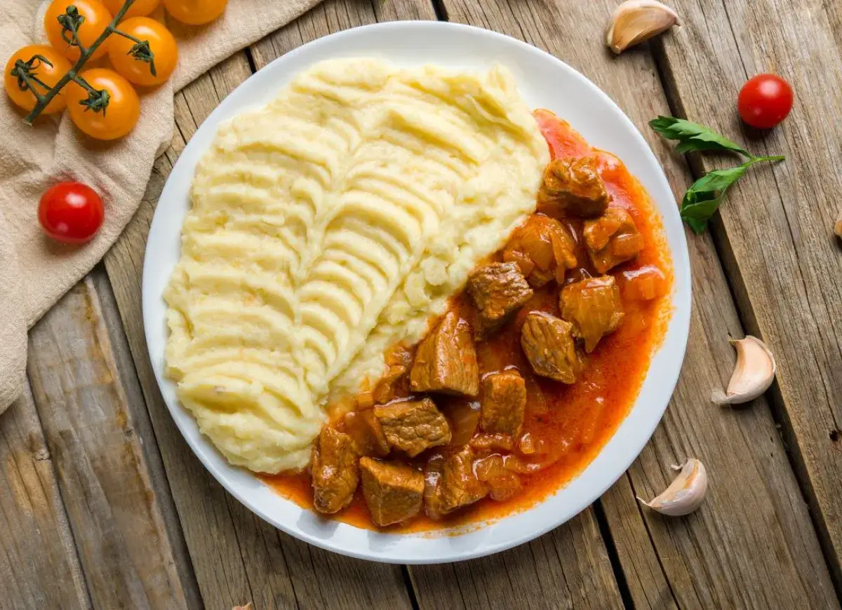 Tradycyjny gulasz wieprzowy z gładkim purée ziemniaczanym, podany na talerzu z pomidorkami i czosnkiem w tle.