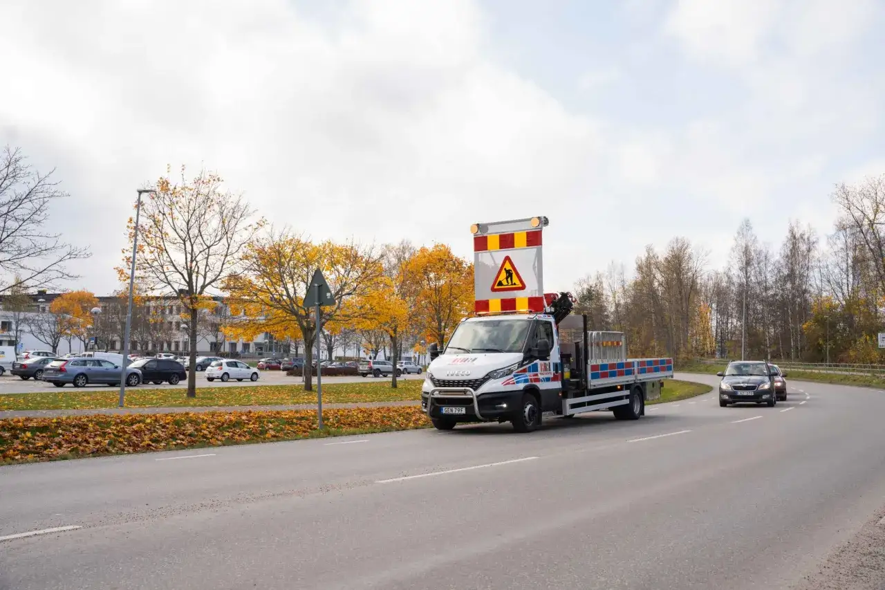 Arbetsfordon med varningsskylt för vägarbete, följer lastplats regler. Höstfärger i bakgrunden.