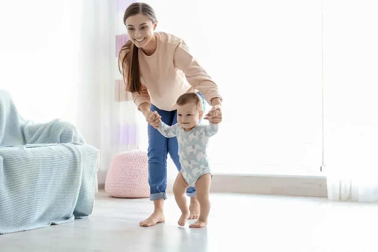 Uśmiechnięta mama pomaga 18-miesięcznemu dziecku, które nie chodzi samodzielnie, stawiać pierwsze kroki.
