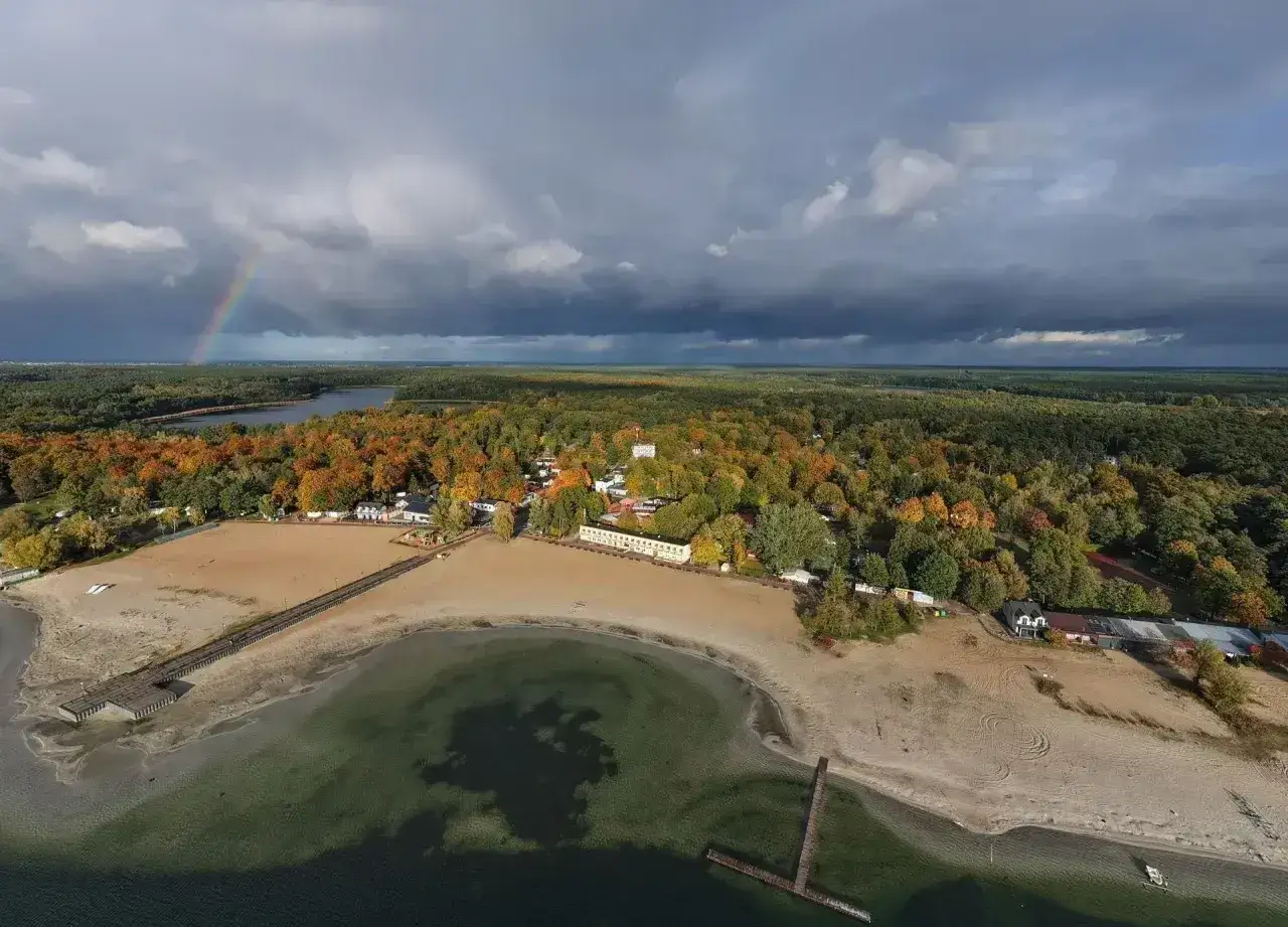 Jesioro Niedzięgiel w jesiennej odsłonie. Tęczowy łuk nad ciemnymi chmurami, piaszczysta plaża i kolorowe drzewa tworzą malowniczy krajobraz.
