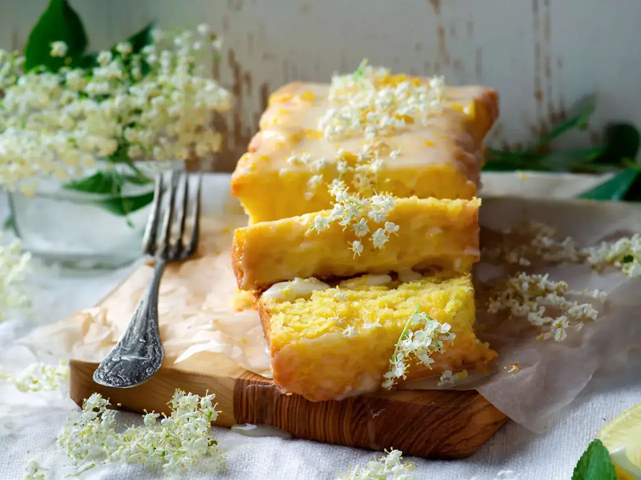 Saftiger holunder kuchen mit Zitronenglasur und frischen Holunderblüten verziert, daneben eine Gabel.