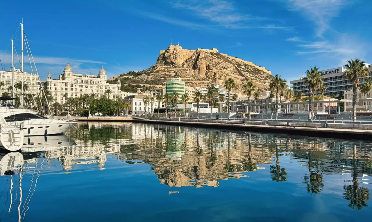 Widok na port w prowincji Alicante z zamkiem Santa Bárbara na wzgórzu i odbiciem budynków w spokojnej wodzie.