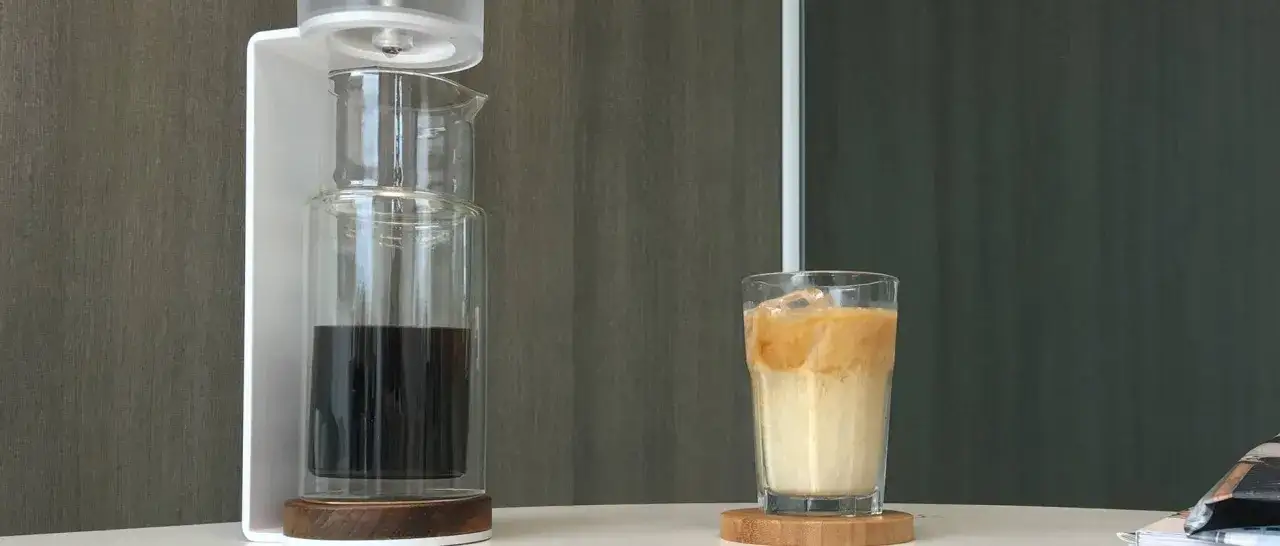 Un bicchiere di **cold brew latte** con ghiaccio, accanto a un infusore per caff&egrave;.