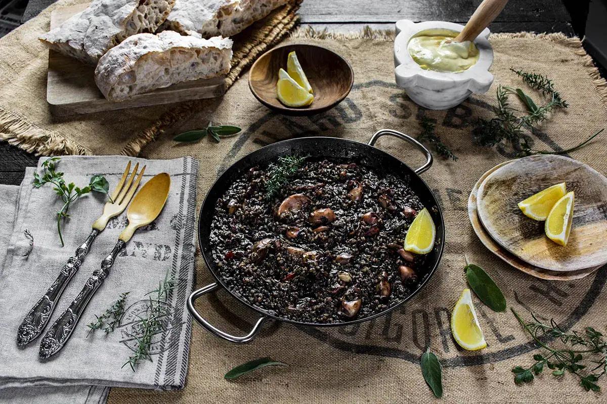 Arroz negro con calamares, servido en paellera, acompañado de pan rústico, limón y alioli.