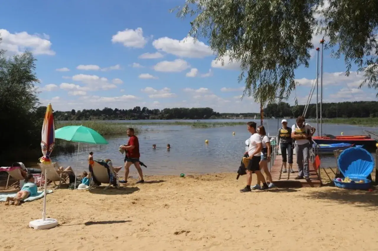 Słoneczny dzień nad zalewem przeczycko-siewierskim. Ludzie odpoczywają na plaży, niektórzy kąpią się w wodzie, inni przygotowują się do rejsu.