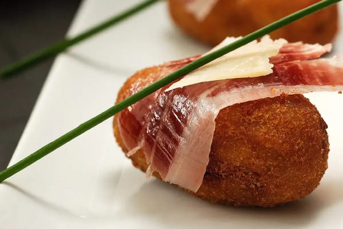 Croquetas de jam&oacute;n, doradas y crujientes, envueltas en jam&oacute;n serrano y coronadas con una lasca de queso.