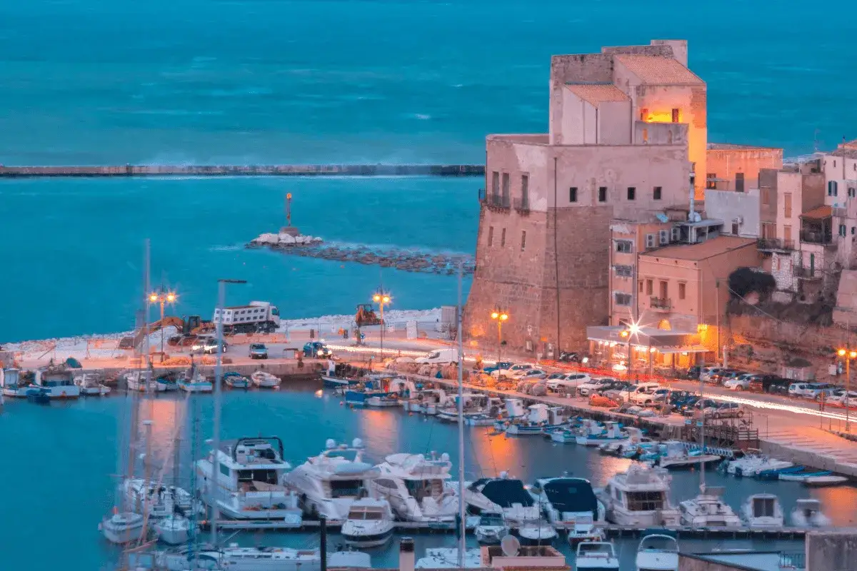 Porto di Castellammare del Golfo al crepuscolo, con barche ormeggiate e un castello antico illuminato.