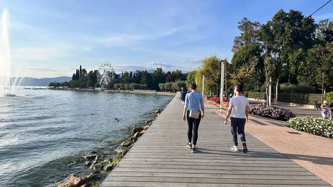 Passeggiata sul lungolago di Cisano di Bardolino, con fontana, ruota panoramica e gente che cammina.