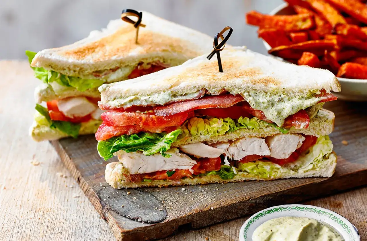 Zwei saftige Club Sandwiches mit Hähnchen, Speck, Tomaten und Salat auf einem Holzbrett, daneben Süßkartoffel-Pommes.