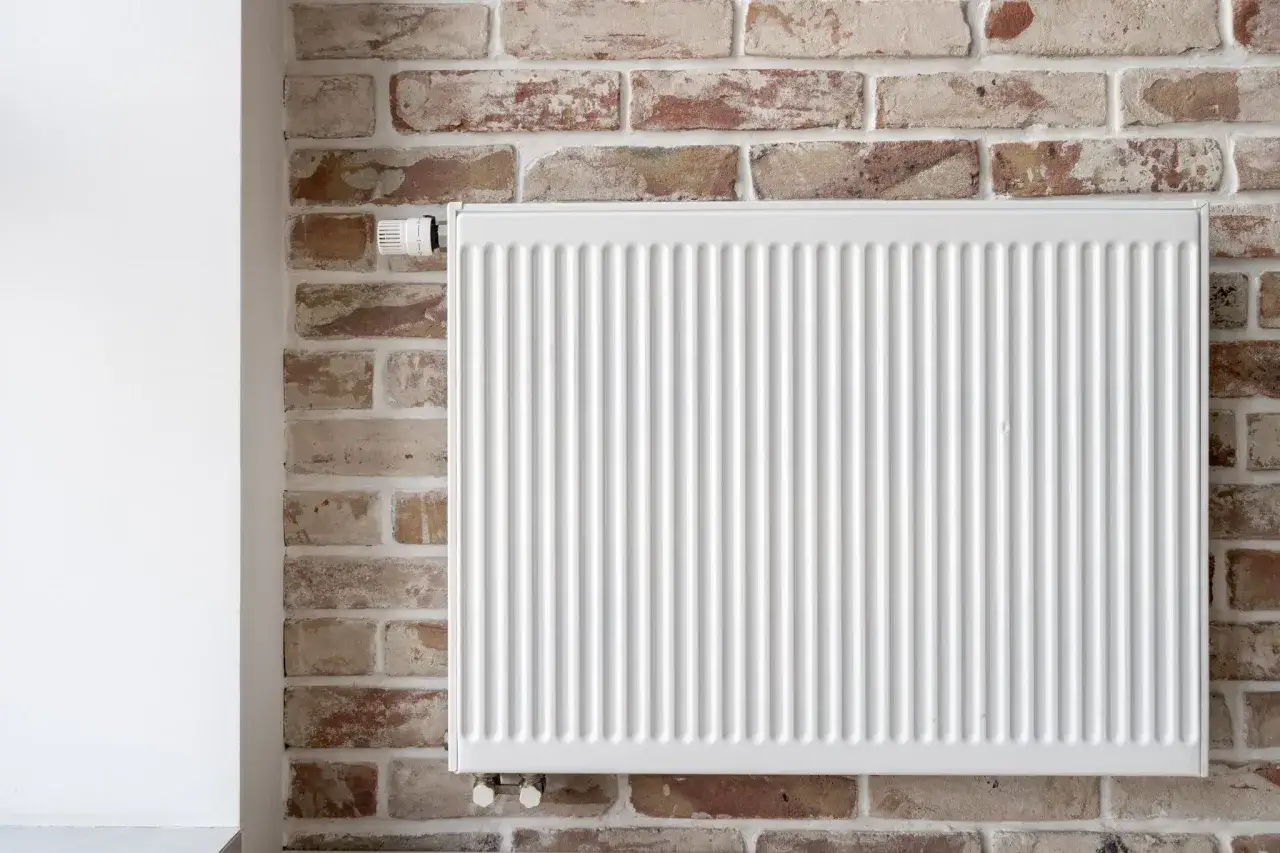 Un radiateur blanc est fixé sur un mur de briques apparentes. Le thermostat est visible sur le côté gauche. Ce mur chauffant assure une ambiance chaleureuse.