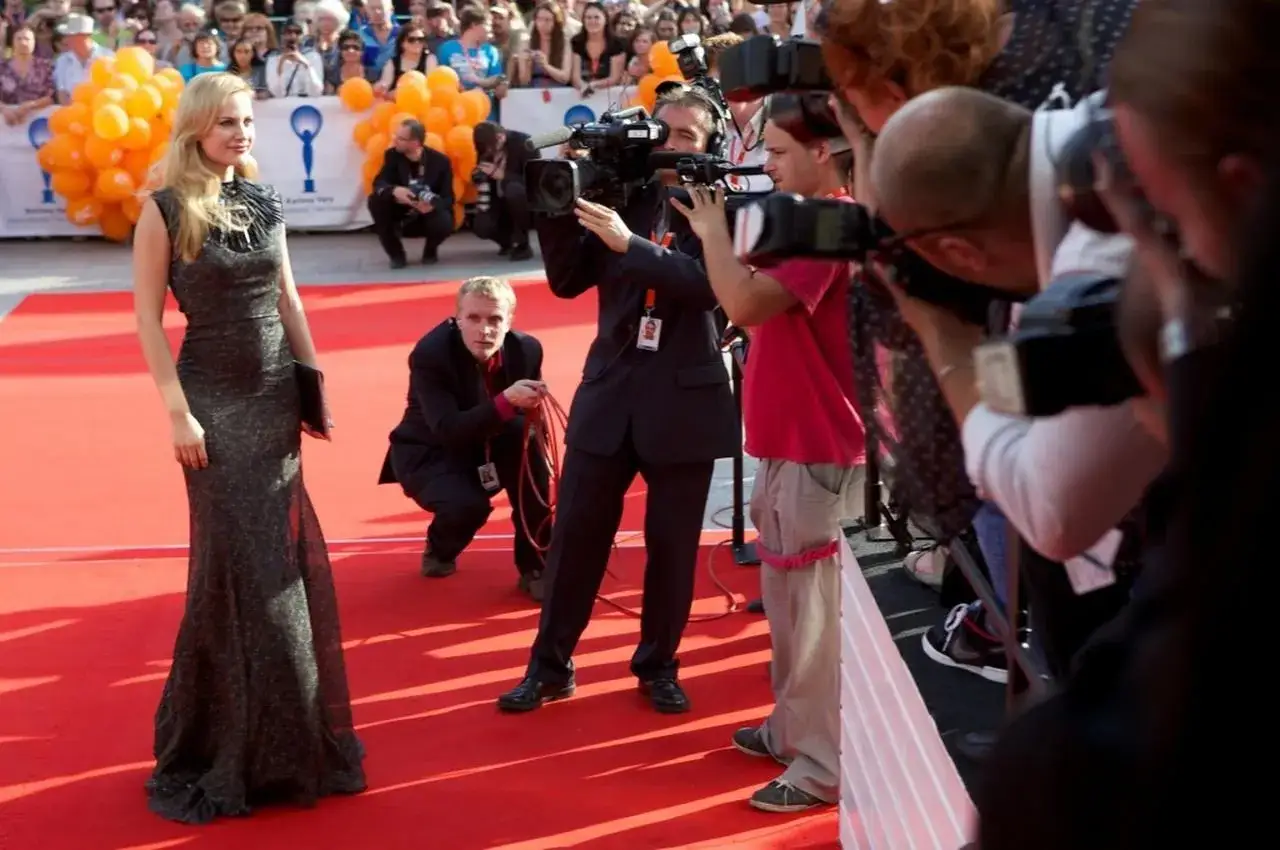 Glamour auf dem roten Teppich beim Karlovy Vary Film Festival. Eine Frau in einem eleganten Kleid posiert, w&auml;hrend Kameraleute und Fotografen sie umringen.