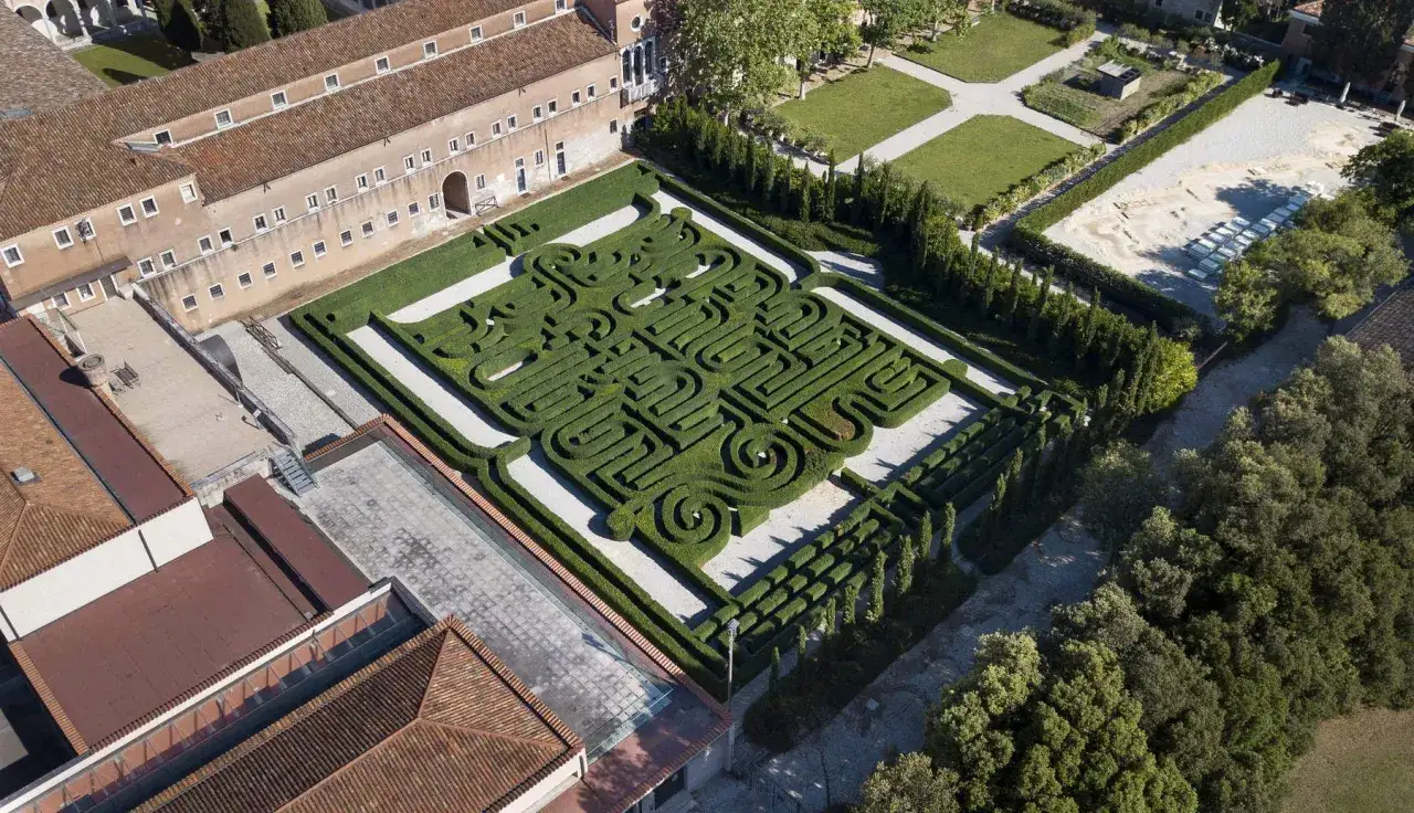 Un giardino con un intricato labirinto di siepi, che ricorda un labirinto borges, circondato da edifici storici e alberi lussureggianti.