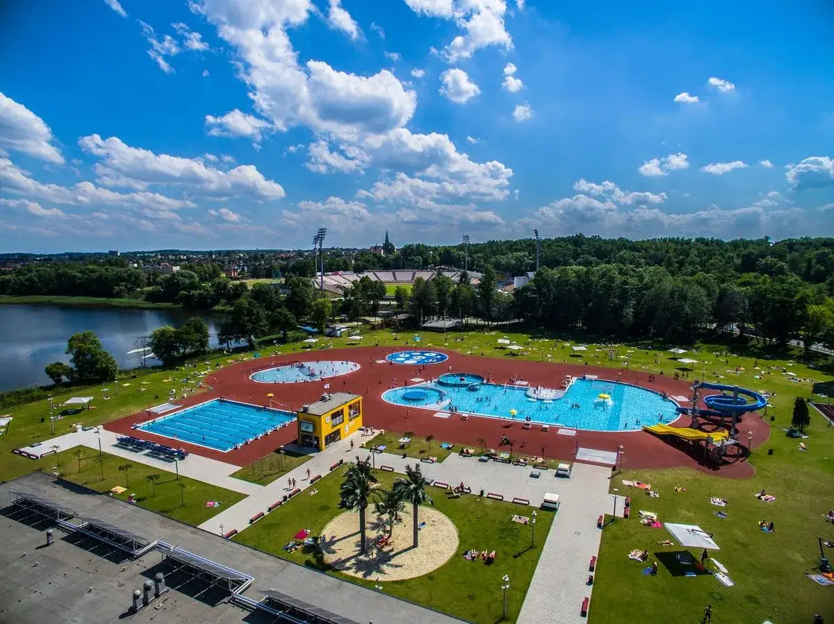 Letni dzień nad wodą. Widać rozległy kompleks basen&oacute;w z wodnymi atrakcjami, zjeżdżalniami i leżakami. W tle jezioro i stadion.