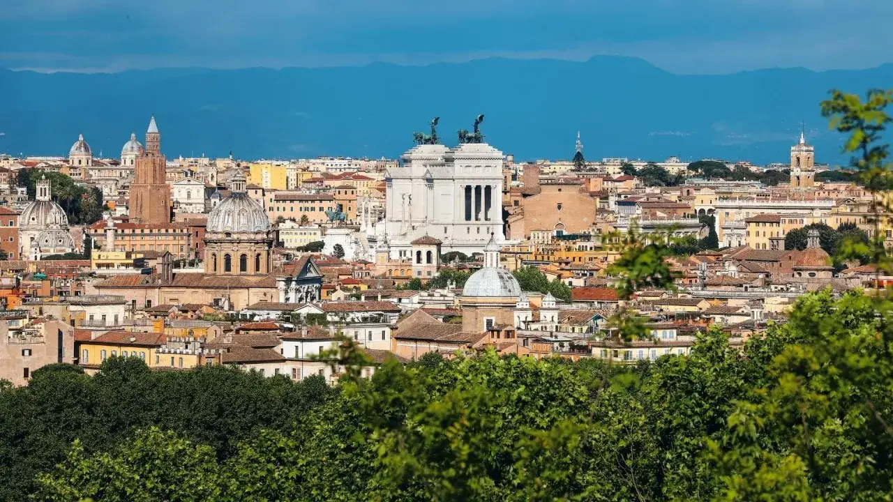 Panorama di Roma 24h: cupole, monumenti e tetti rossi sotto un cielo azzurro, con montagne sullo sfondo.