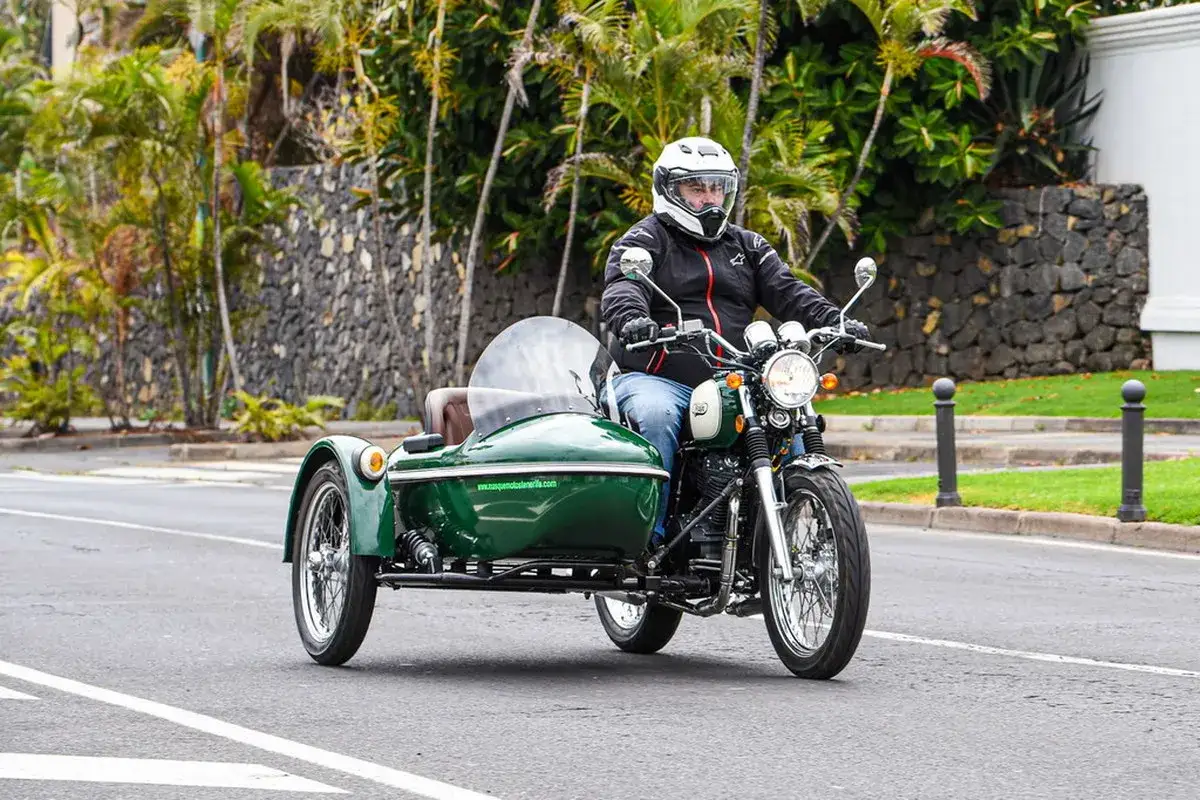 Hombre conduce una moto con sidecar verde por una carretera tropical.