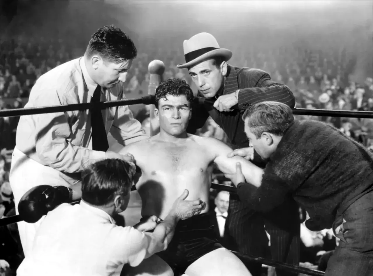 A boxer sits in his corner, attended by his corner man and trainer, while a man in a fedora looks on intently.