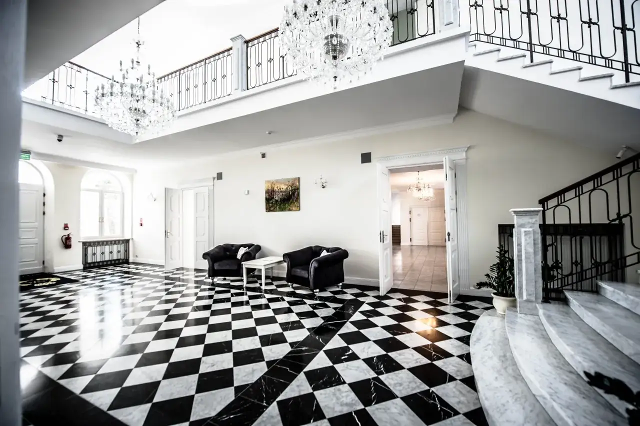 Eleganckie wnętrze hotelu Sobienie Królewskie z kryształowymi żyrandolami, czarno-białą szachownicą na podłodze i marmurowymi schodami.