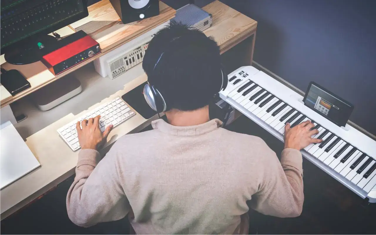 Mann mit Kopfhörern spielt auf einem weißen Keyboard und schreibt eigene Songs. Am Schreibtisch sind Computer, Lautsprecher und ein rotes Audio-Interface.