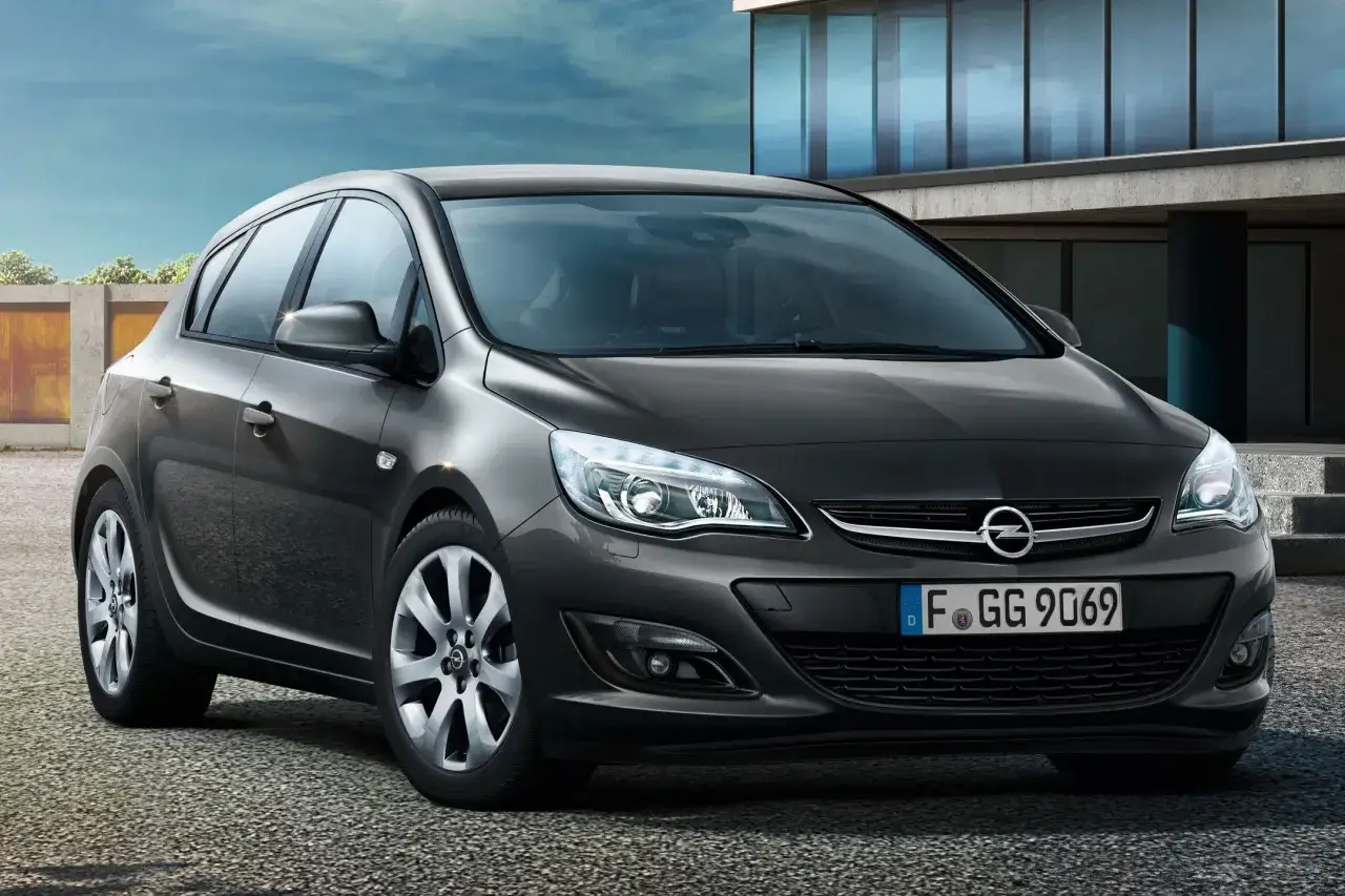Ciemnoszary Opel Astra J z niemieckimi tablicami rejestracyjnymi stoi przed nowoczesnym budynkiem.
