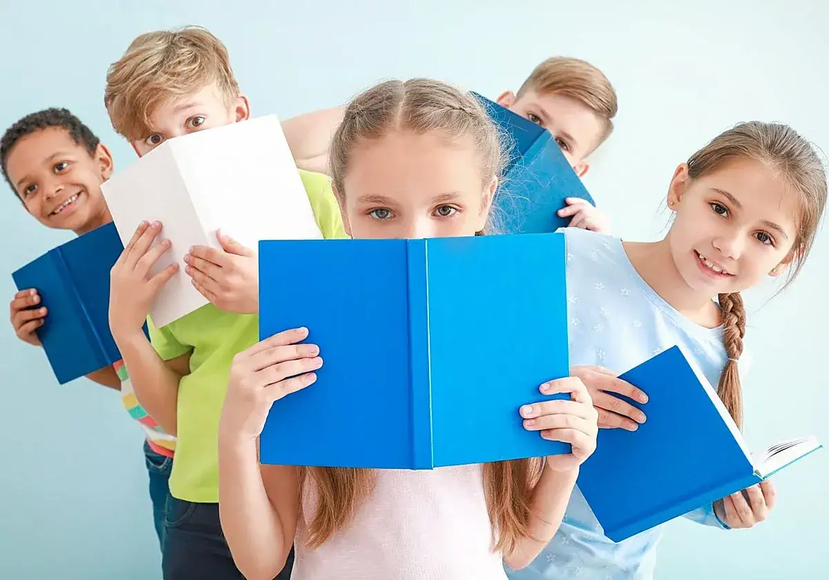 Niños de primaria disfrutan de la lectura comprensiva, escondiéndose detrás de libros azules y blancos.