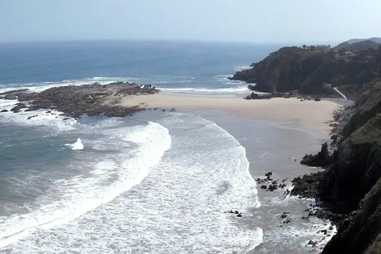 Vista aérea de la **playa de Pechón**, con olas rompiendo en la arena dorada y acantilados verdes.