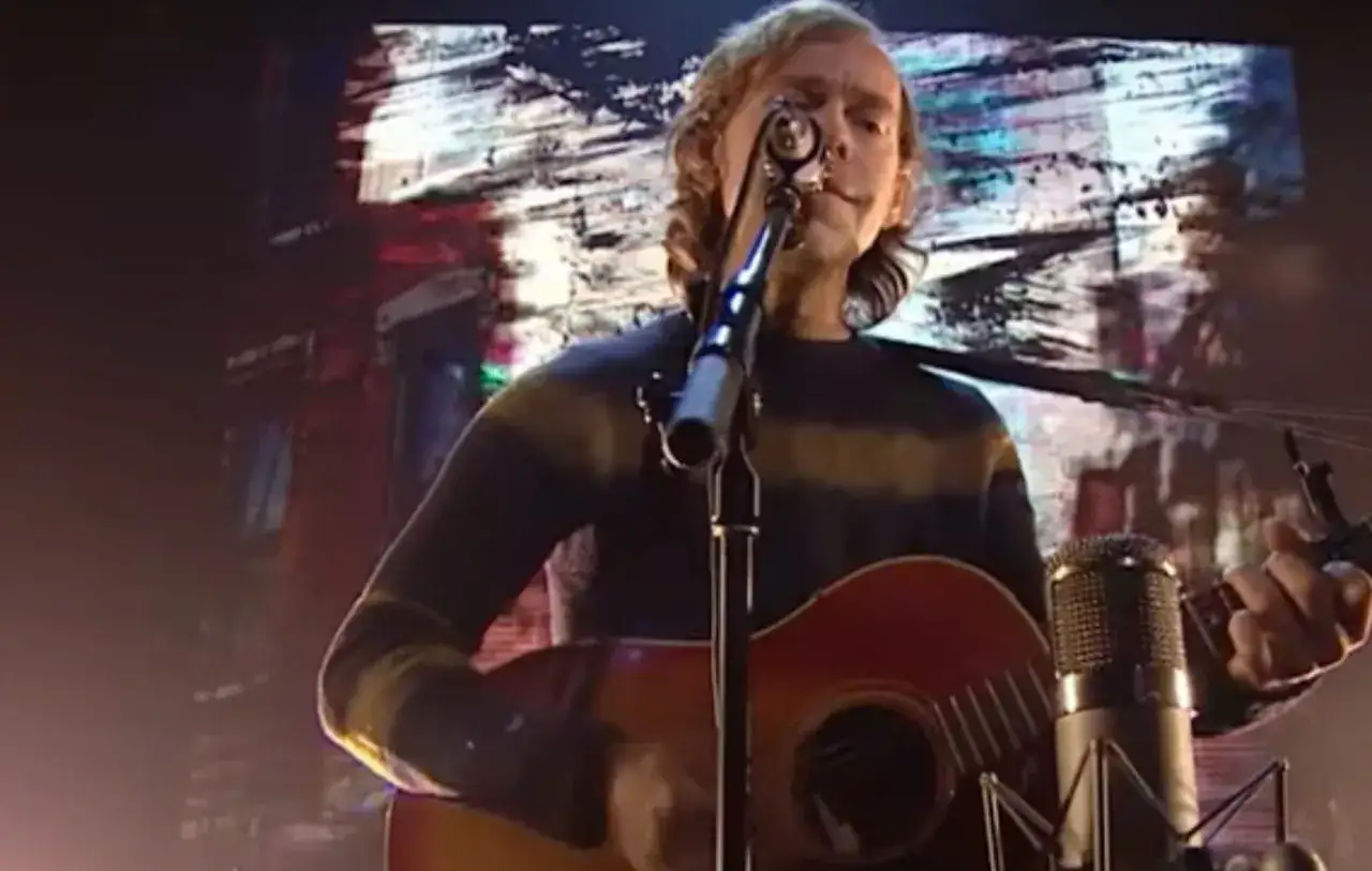 Aaron Dessner performs one of his evocative songs, strumming an acoustic guitar in front of a vibrant, abstract backdrop.