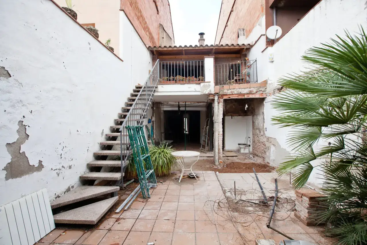 Patio de una casa antigua en proceso de reforma. Escaleras metálicas, escombros y una palmera anuncian el inicio de la transformación.