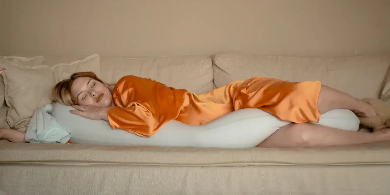 En kvinna sover p&aring; en soffa med en stor kudde mellan benen. Hon har en orange satinpyjamas p&aring; sig. Vilken sida ska man sova p&aring;?