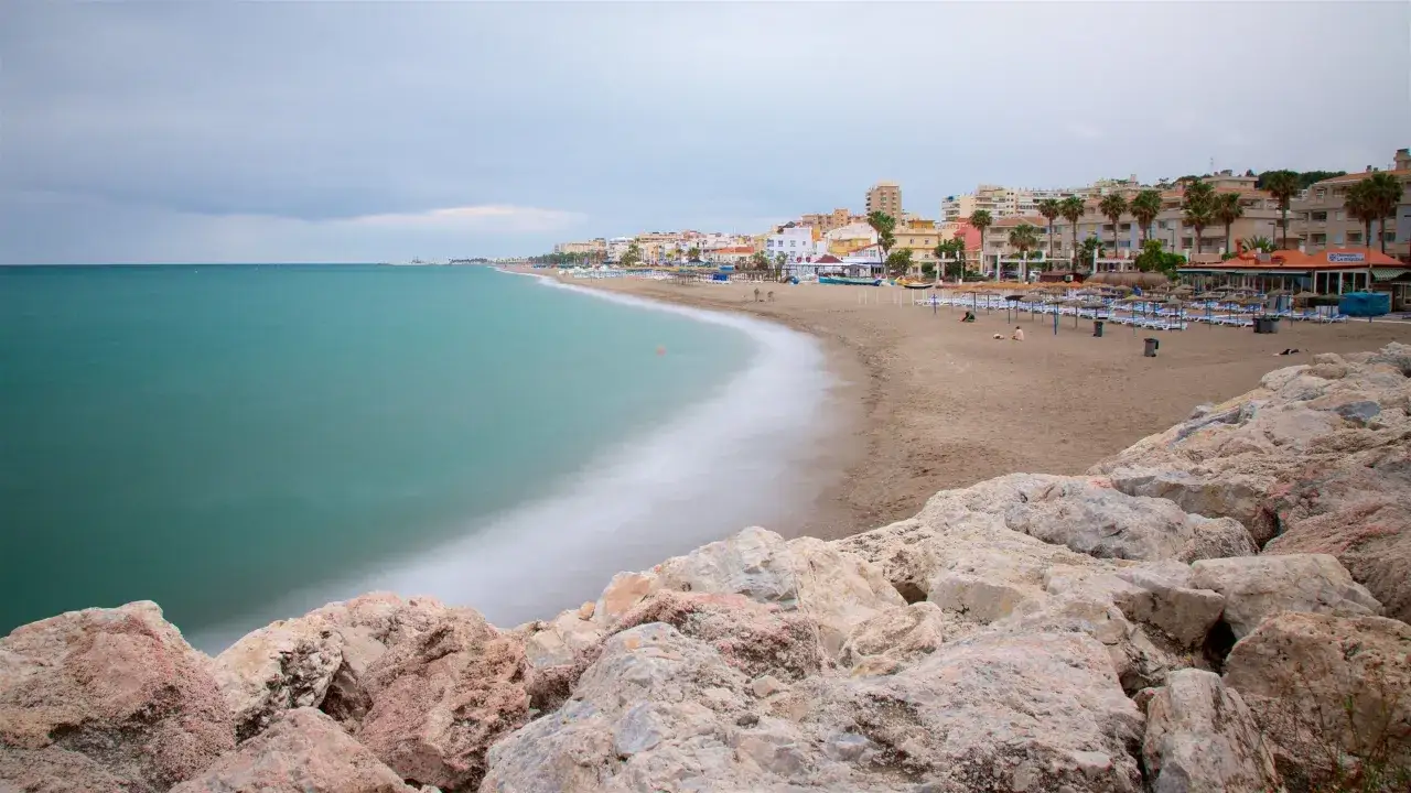 T&uuml;rkisblaues Meer trifft auf den Sandstrand der playa de la carihuela. Felsen im Vordergrund, dahinter eine Stadtkulisse mit Palmen.