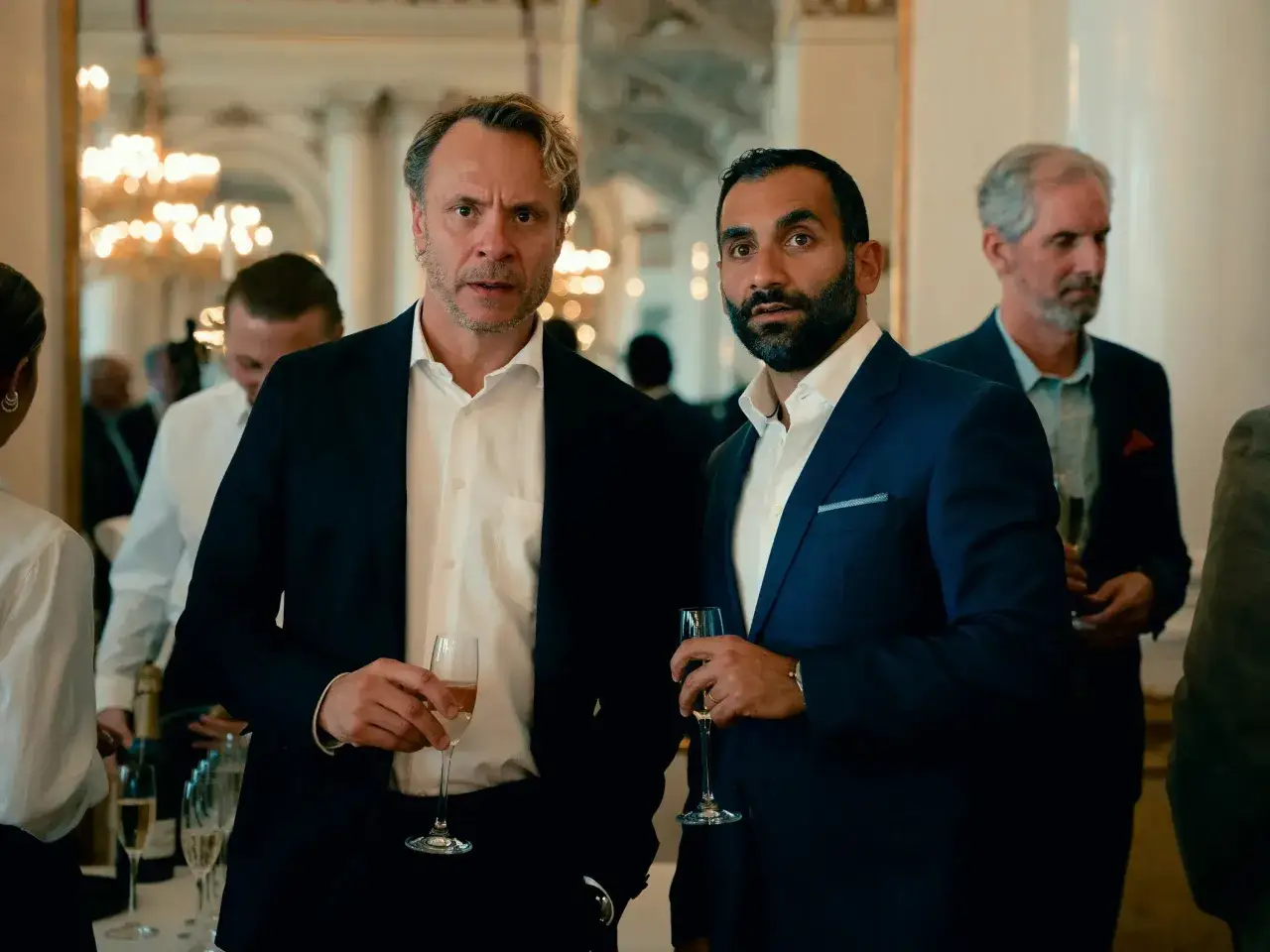 Två män i kostym med champagneglas i handen, kanske en scen från hundarna serie.