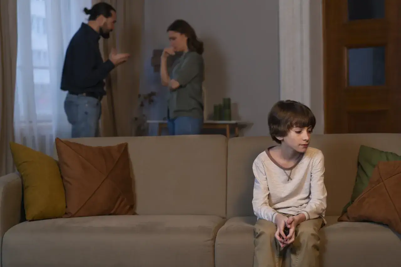 Un enfant seul sur un canapé, le regard perdu, tandis que ses parents se disputent en arrière-plan. Il se sent mal aimé.