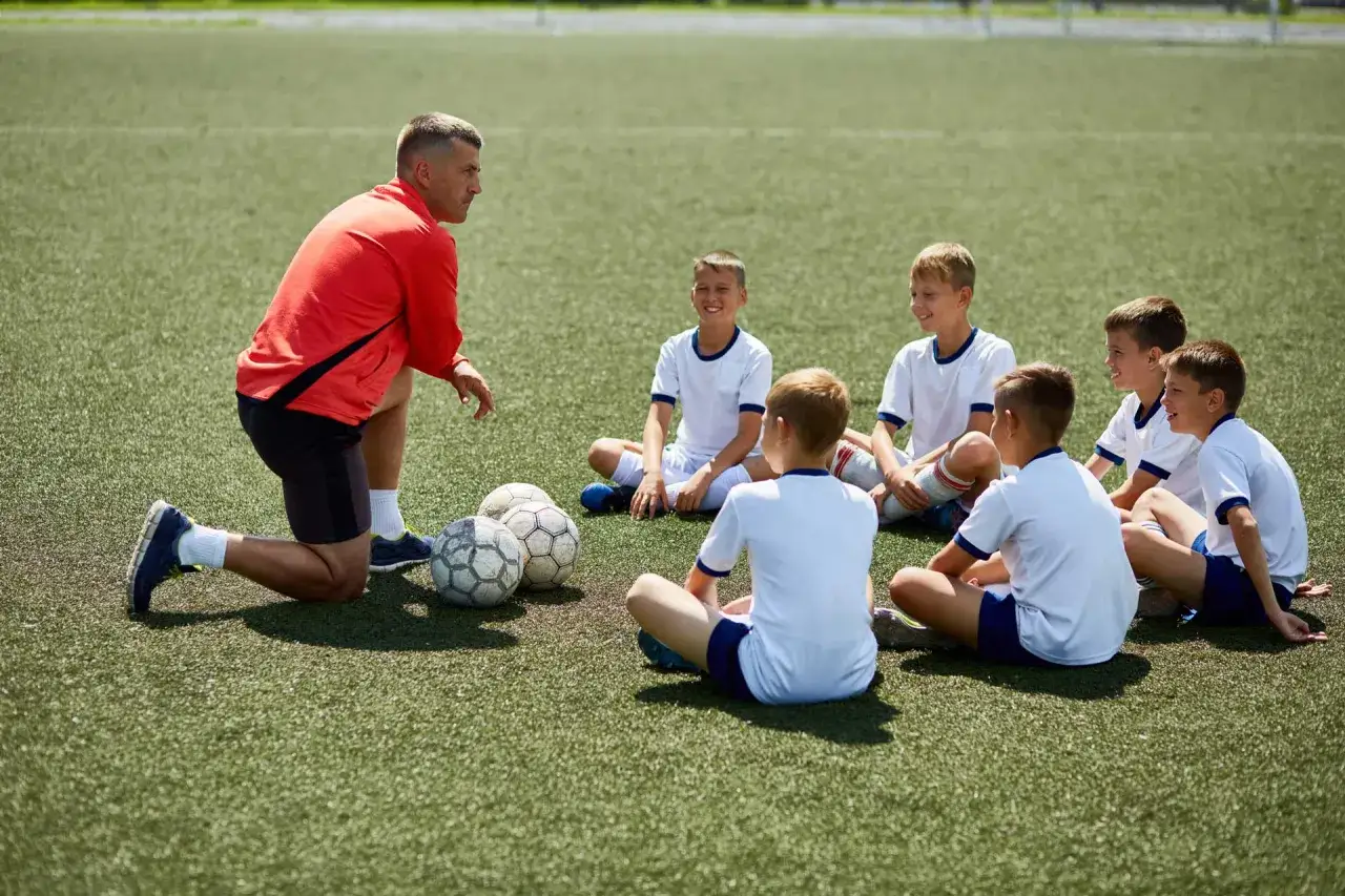 Trener prowadzi kurs trenera piłki nożnej dla dzieci. Młodzi piłkarze siedzą na boisku, słuchając jego wskazówek.