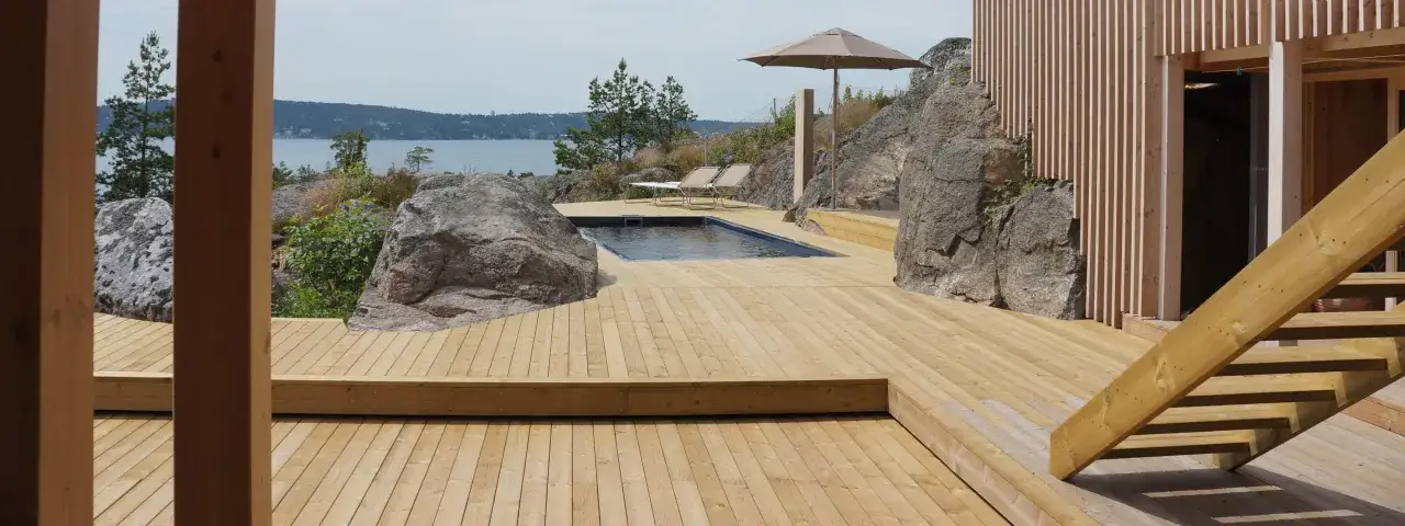 En stor altan i trä med en pool och solstolar, omgiven av klippor och med utsikt över vattnet. Perfekt för en fris trall.