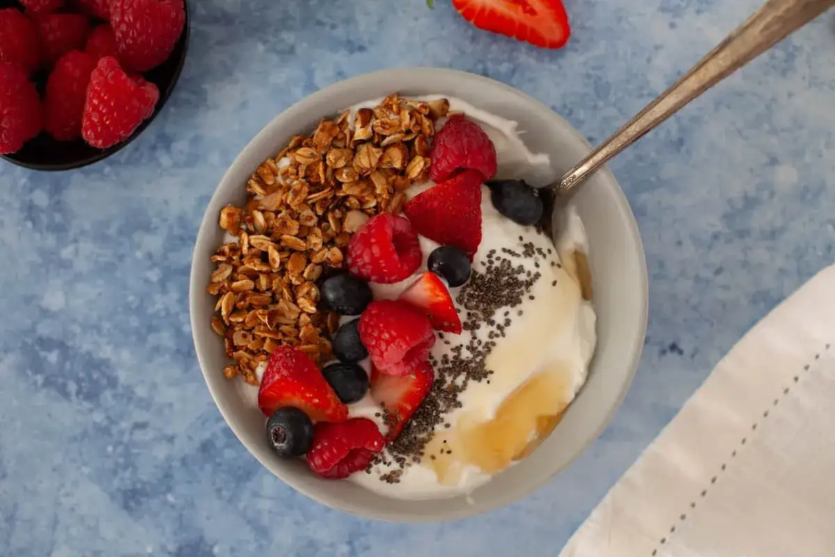 Schüssel mit Joghurt, Granola, Beeren und Honig. Leinsamen sind über den Joghurt gestreut.