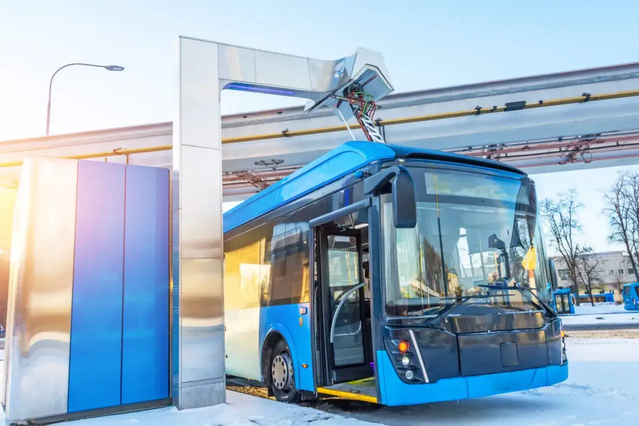 Nowoczesny, niebieski autobus elektryczny podłączony do ładowarki na tle zimowego krajobrazu.