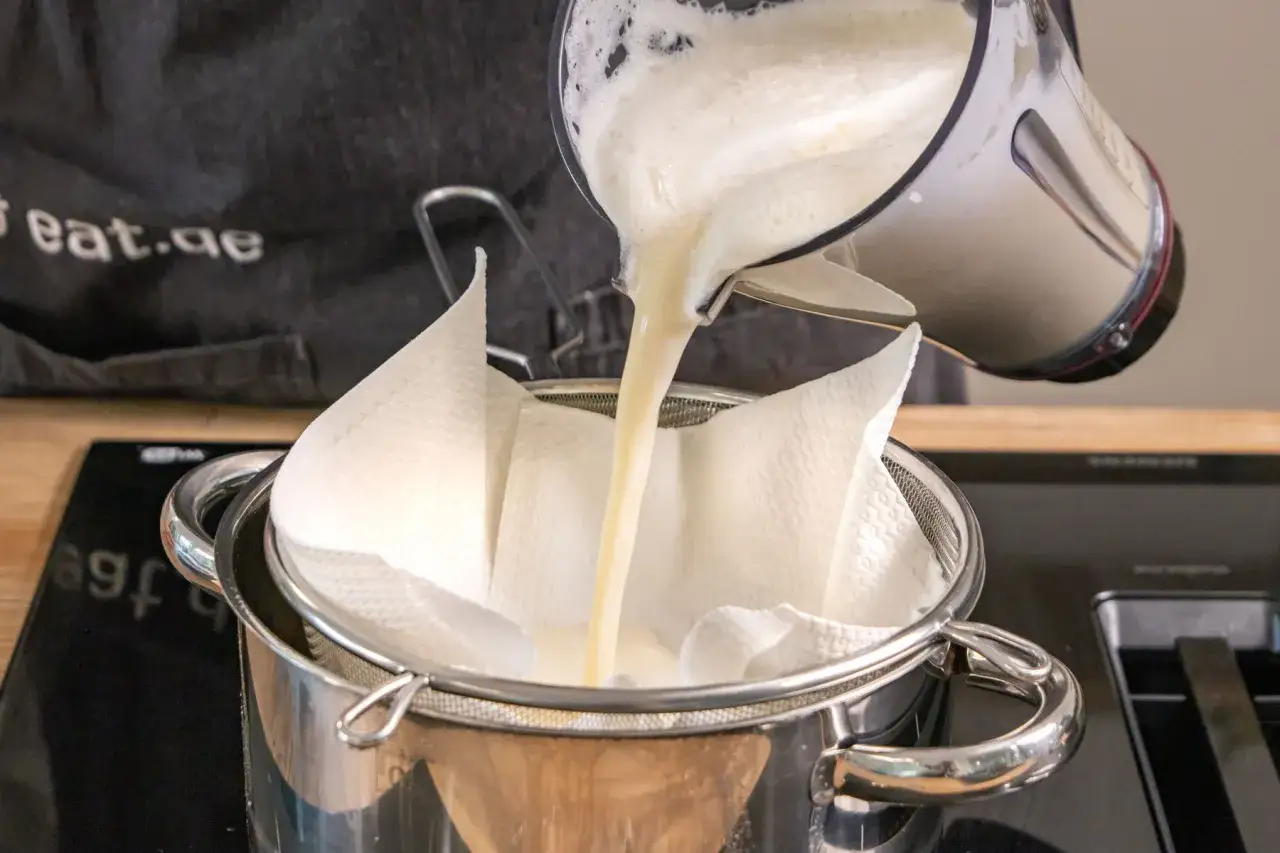 Fl&uuml;ssigkeit aus einem Mixer wird durch ein Sieb mit K&uuml;chenpapier gegossen, um **sojamilch selber machen** zu erleichtern.