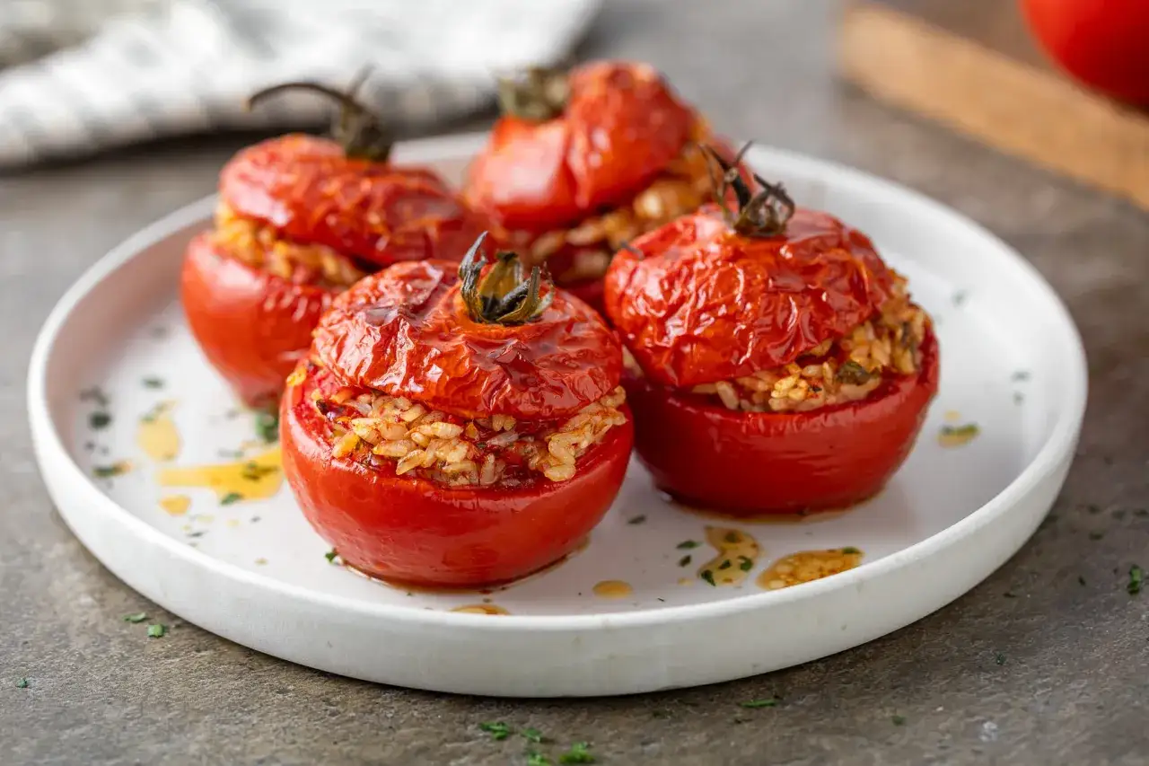 Quattro pomodori ripieni di riso, cotti al forno, serviti su un piatto bianco con erbe aromatiche e olio.
