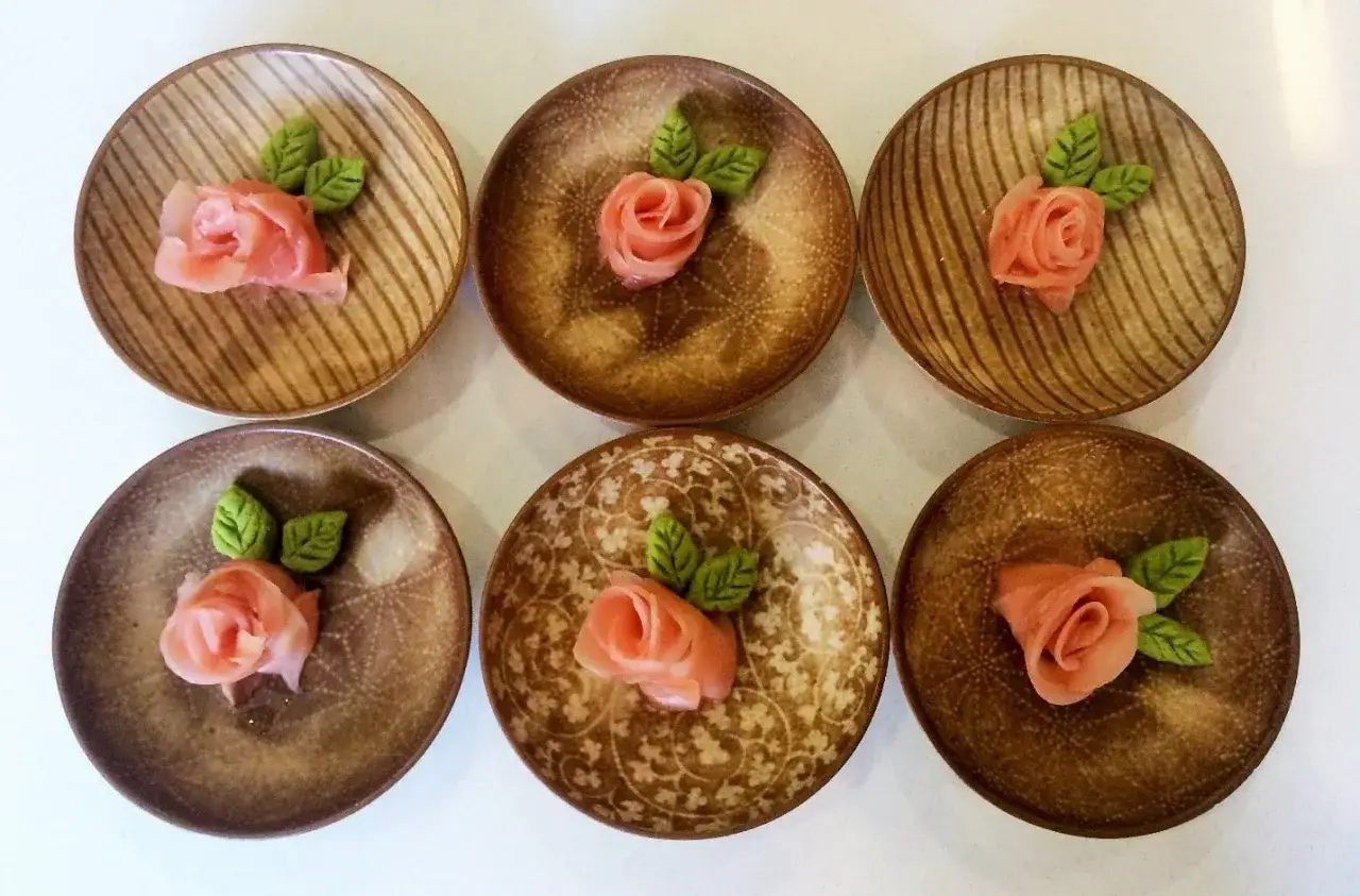 Sześć talerzyków z różami z marynowanego imbiru do sushi, ozdobione zielonymi listkami.