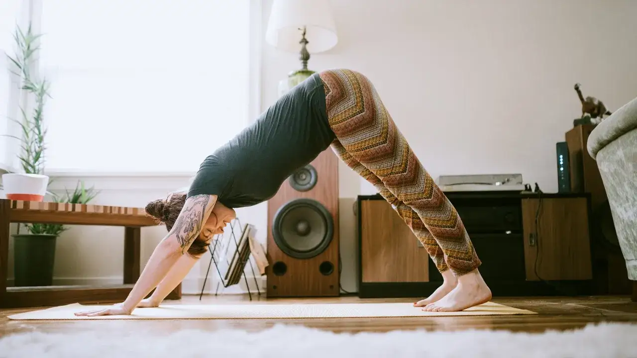 Kvinna i m&ouml;nstrade byxor g&ouml;r en ned&aring;tg&aring;ende hund-position, en del av tibetansk yoga, p&aring; en gul matta i ett vardagsrum.