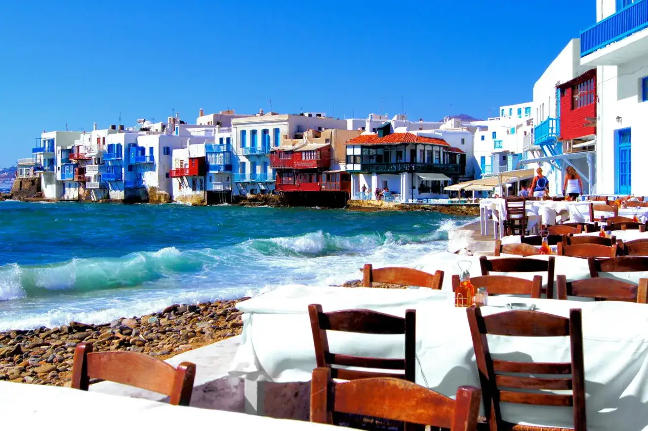 Cosa vedere a Mykonos in crociera: case bianche con balconi blu sul mare, tavoli apparecchiati per un pranzo vista onde.