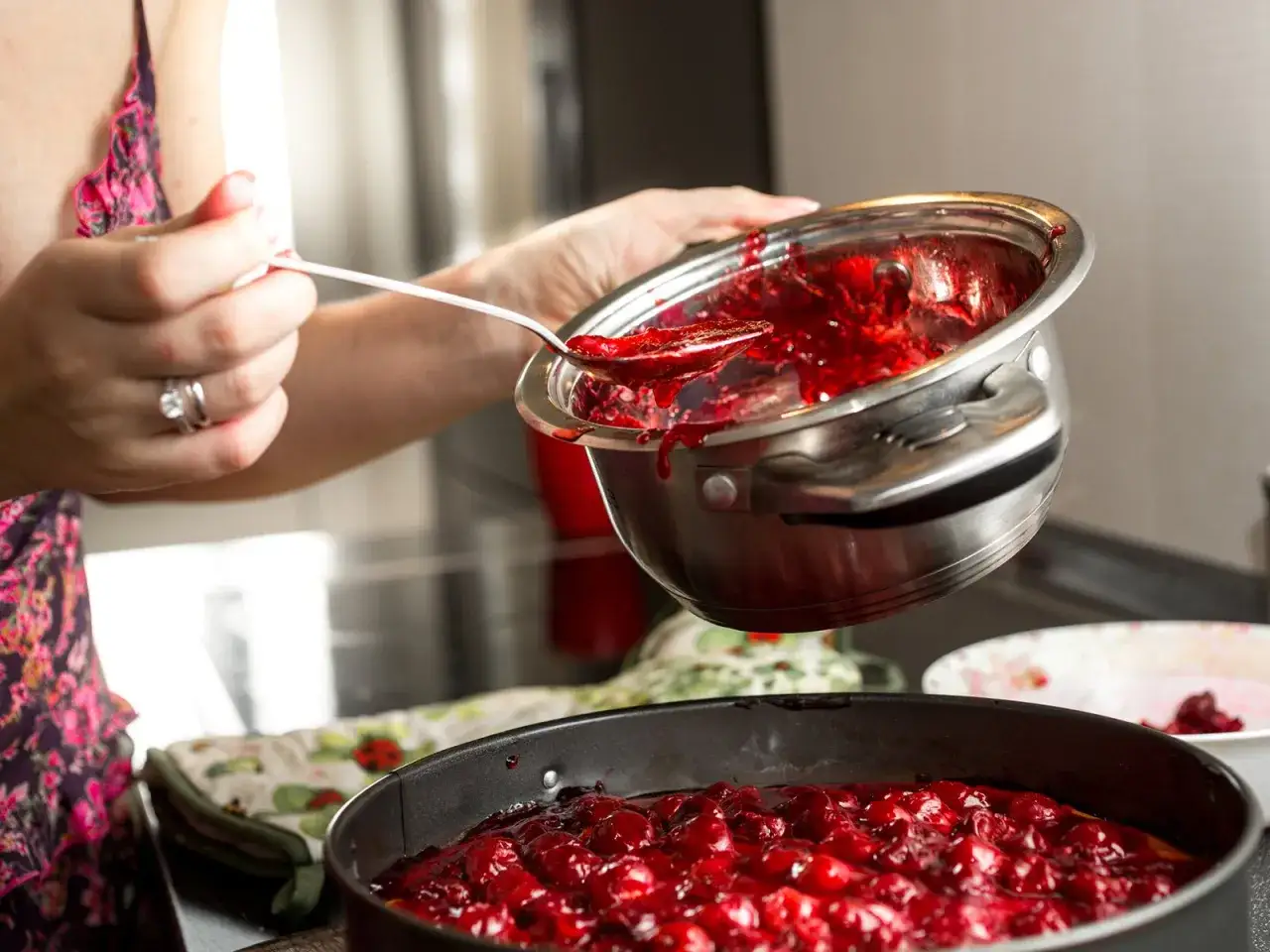 Eine Person sch&ouml;pft mit einem L&ouml;ffel fl&uuml;ssige Kirschmasse aus einem Topf in eine Kuchenform. Die Marmelade ist noch nicht fest geworden.