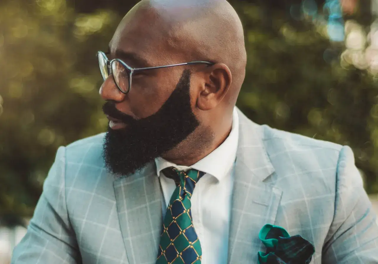 Un homme chauve porte un costume &agrave; carreaux gris, une chemise blanche et une cravate verte et bleue. Sa barbe mouche est taill&eacute;e avec pr&eacute;cision.