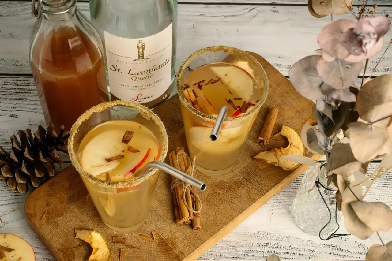 Zwei Gl&auml;ser mit Apfel-Zimt-Wasser, garniert mit Apfelscheiben und Zimtstangen. Daneben eine Flasche Apfelsaft und St. Leonhards Quelle Mineralwasser.