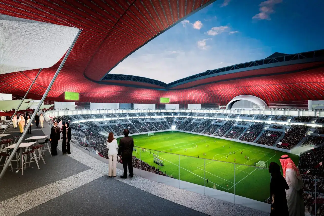 Widok na pełen widz&oacute;w stadion Al Bayt z zieloną murawą i czerwoną, falistą konstrukcją dachu.